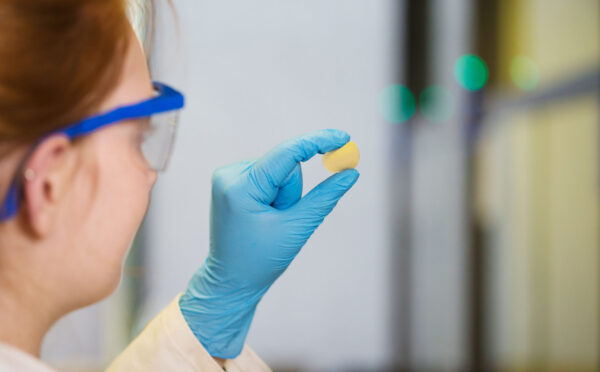 Person wearing safety goggles and blue gloves holds a small yellow object in a lab.